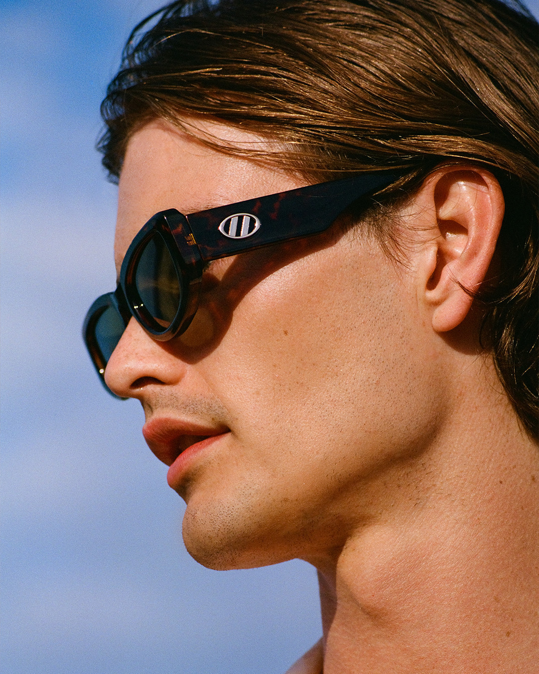 Close-up of a young adult male with brown hair, wearing patterned sunglasses, against a clear blue sky background.