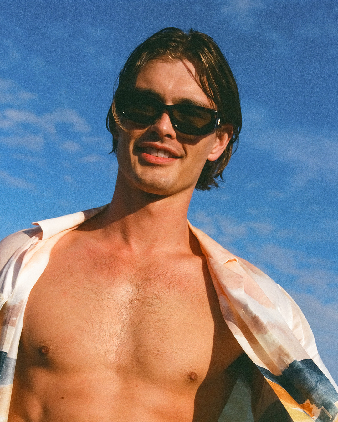Portrait of a shirtless, tan adult male wearing sunglasses against a clear blue sky. He has brown hair and a slight smile.