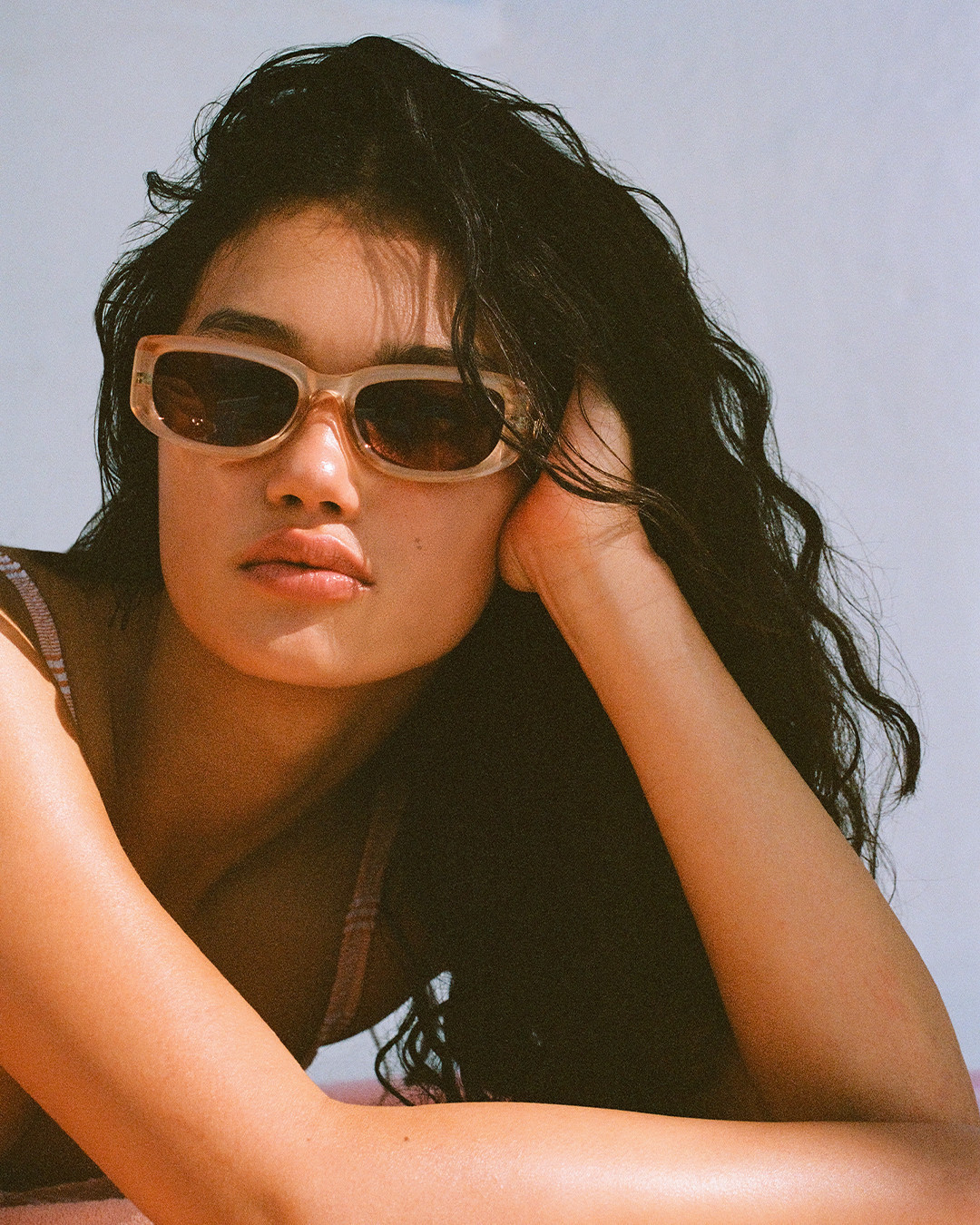 Portrait of a young woman with wavy black hair wearing sunglasses, posing with her hand near her face against a plain background.