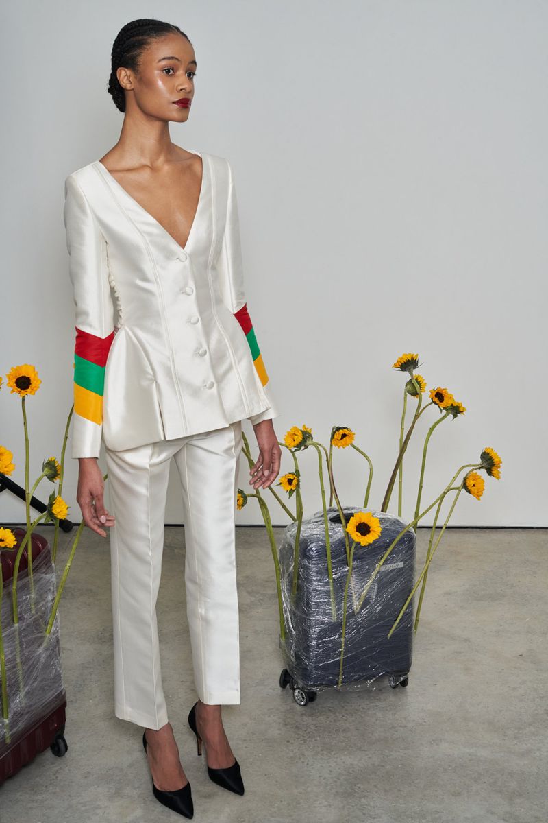 Elegant woman in ivory suit with vibrant sleeve detail poses amidst sunflowers and vintage luggage for high-fashion editorial.