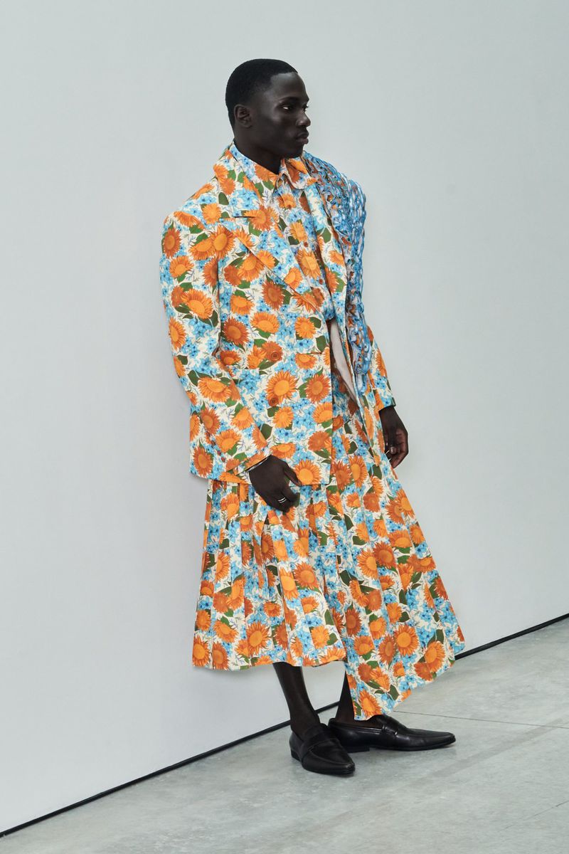 Man wearing a floral suit and skirt posing indoors. The ensemble features a mix of orange and blue flowers over a white background.