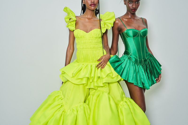 Two dark-skinned women pose in couture fashion: one in long neon dress, the other in a short emerald bustier dress against a white wall.