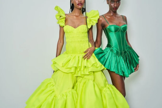Two dark-skinned women pose in couture fashion: one in long neon dress, the other in a short emerald bustier dress against a white wall.