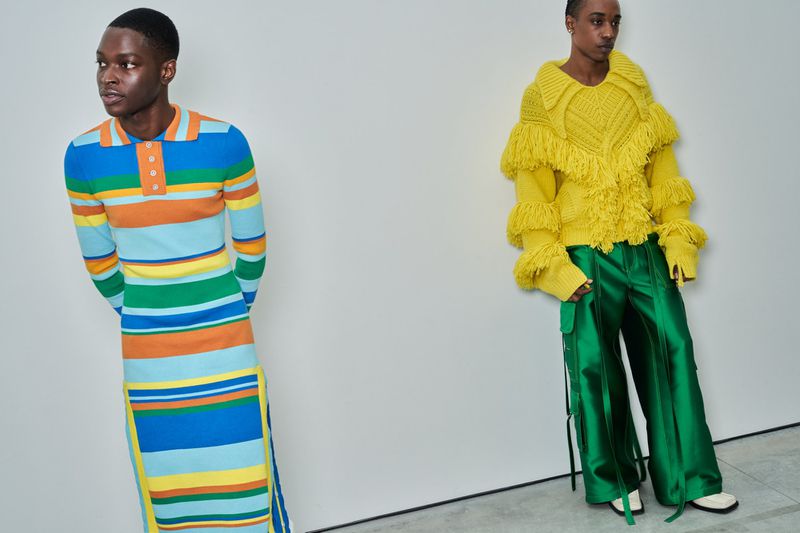 Two people modeling colorful and unique knitwear against a plain white wall backdrop, showcasing bold fashion designs in a studio setting.