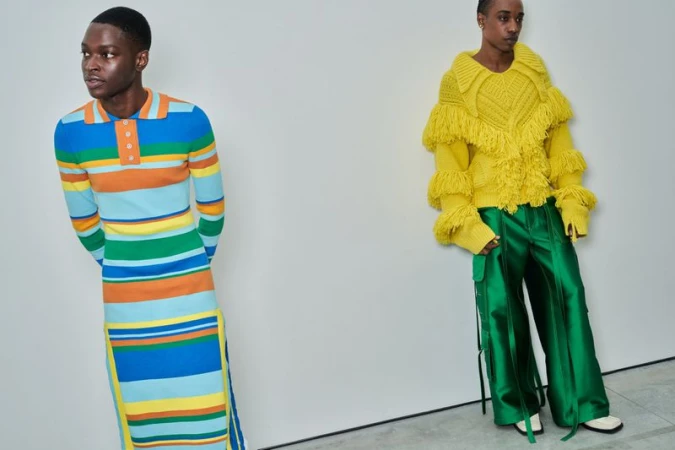 Two people modeling colorful and unique knitwear against a plain white wall backdrop, showcasing bold fashion designs in a studio setting.