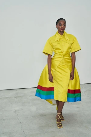 Fashionable woman posing in a vibrant yellow dress with multi-colored accents and matching heels against a stark white studio backdrop.