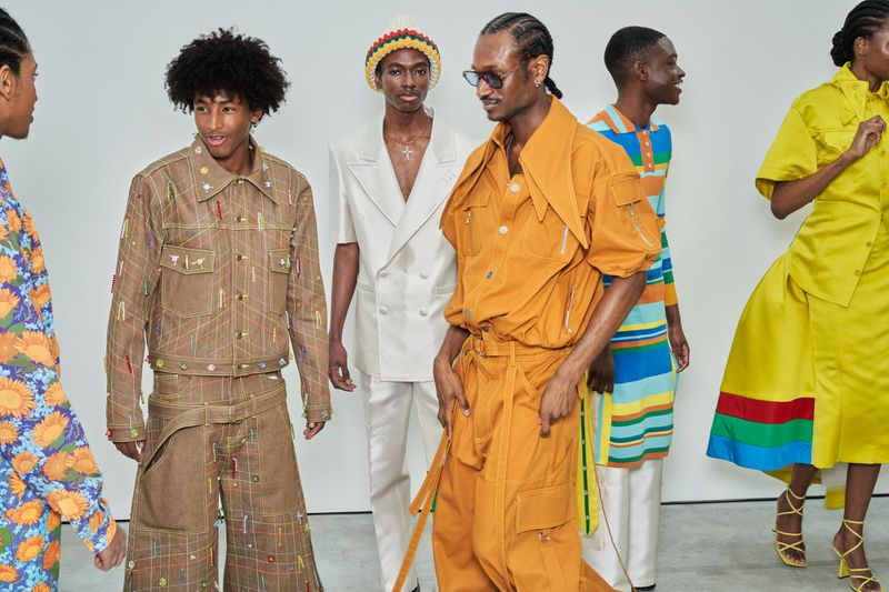 A group of six fashion models are standing in a studio wearing colorful, fashionable clothing; the backdrop is a plain white studio.
