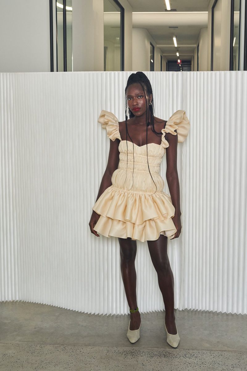 Elegant Black woman in cream puff dress poses in minimalist interior. Shoulder ruffles, tiered skirt, cream pumps. Fashion shoot.