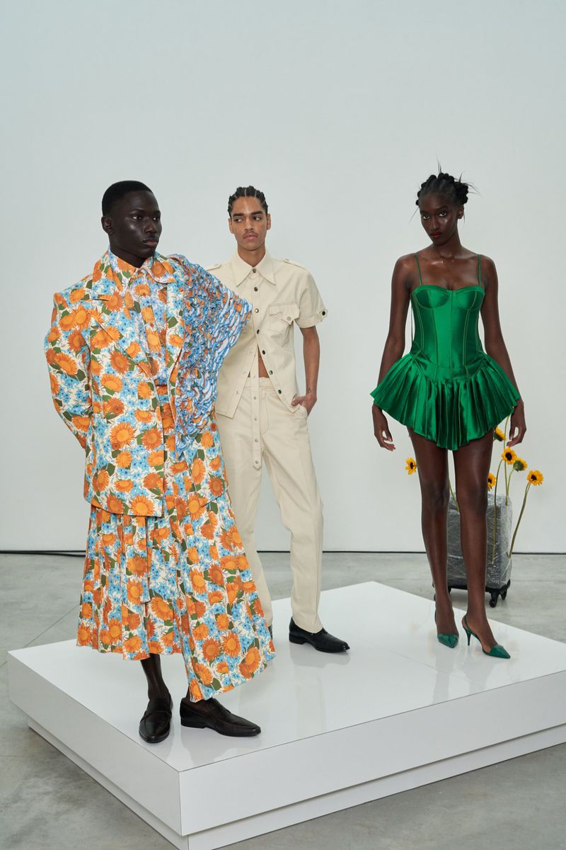 Three fashion models present a new collection on a white platform in a bright studio, featuring vibrant floral prints and tailored designs.