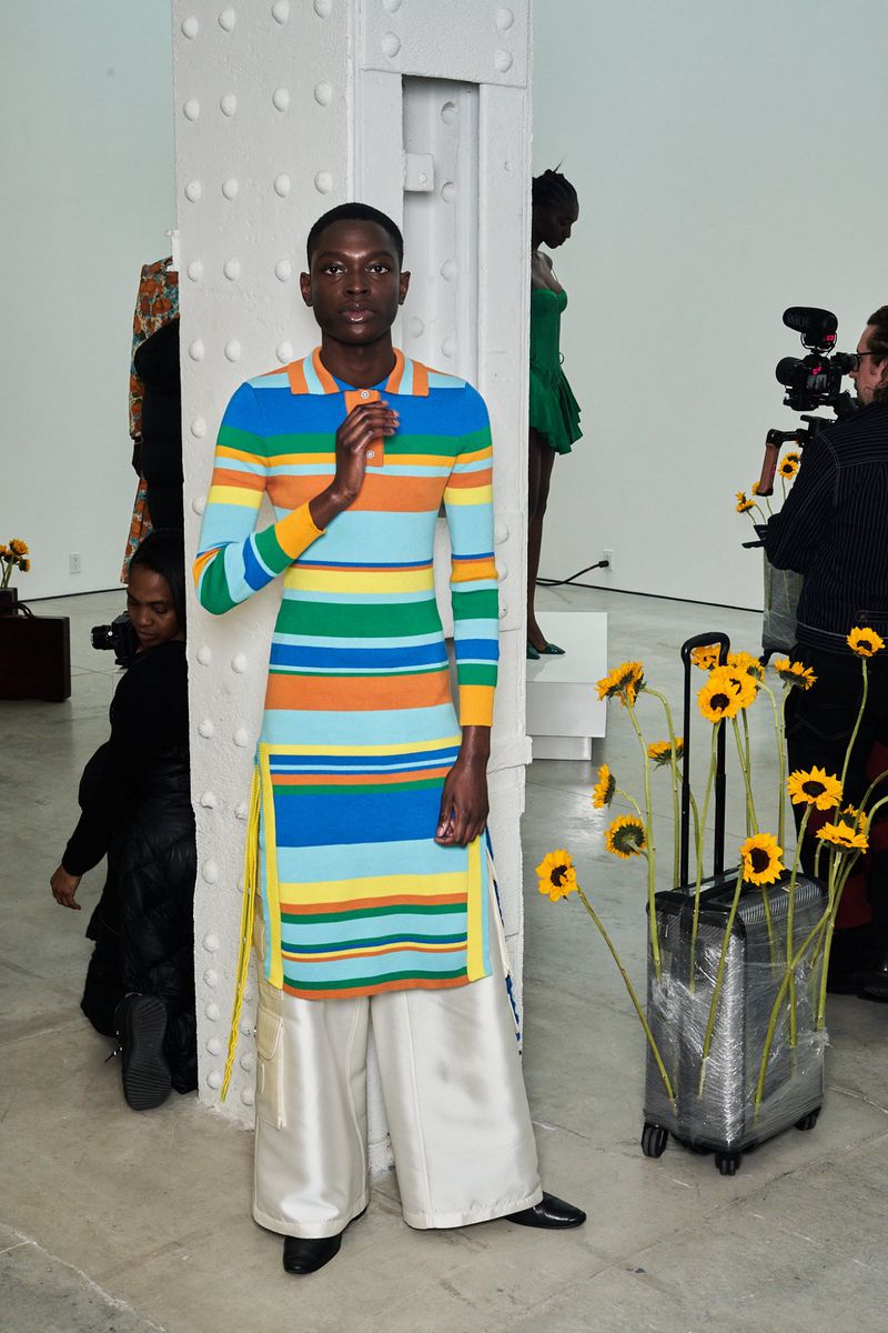 Runway image featuring a Black male model in a colorful striped dress and wide-leg pants standing next to a suitcase of sunflowers.