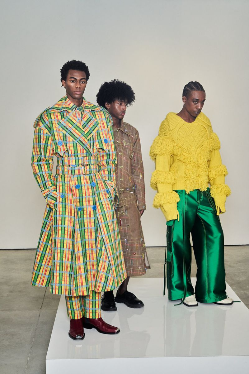 Three fashion models in colorful, patterned and textured clothing stand on a white platform against a neutral background in a studio.