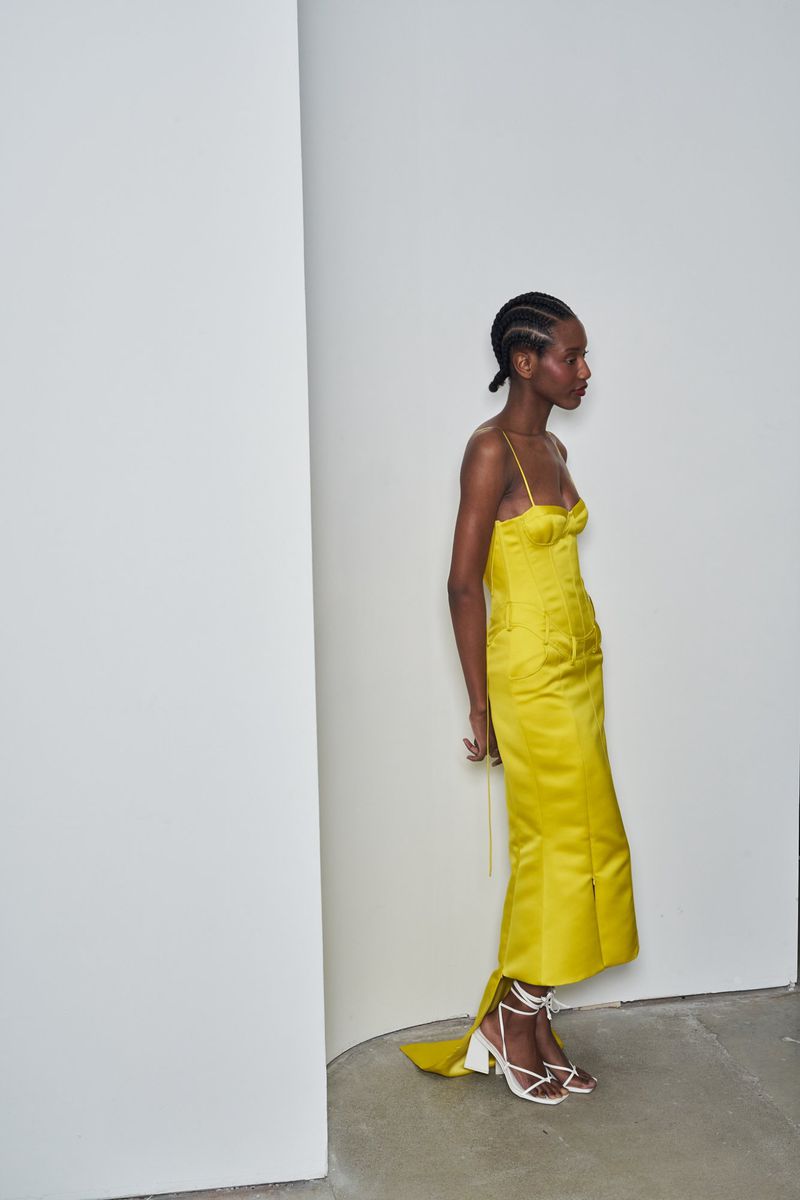 A dark-skinned woman in a yellow dress and white strappy high heels stands in front of a white corner in a fashion shot.