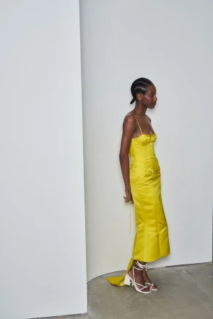 A dark-skinned woman in a yellow dress and white strappy high heels stands in front of a white corner in a fashion shot.