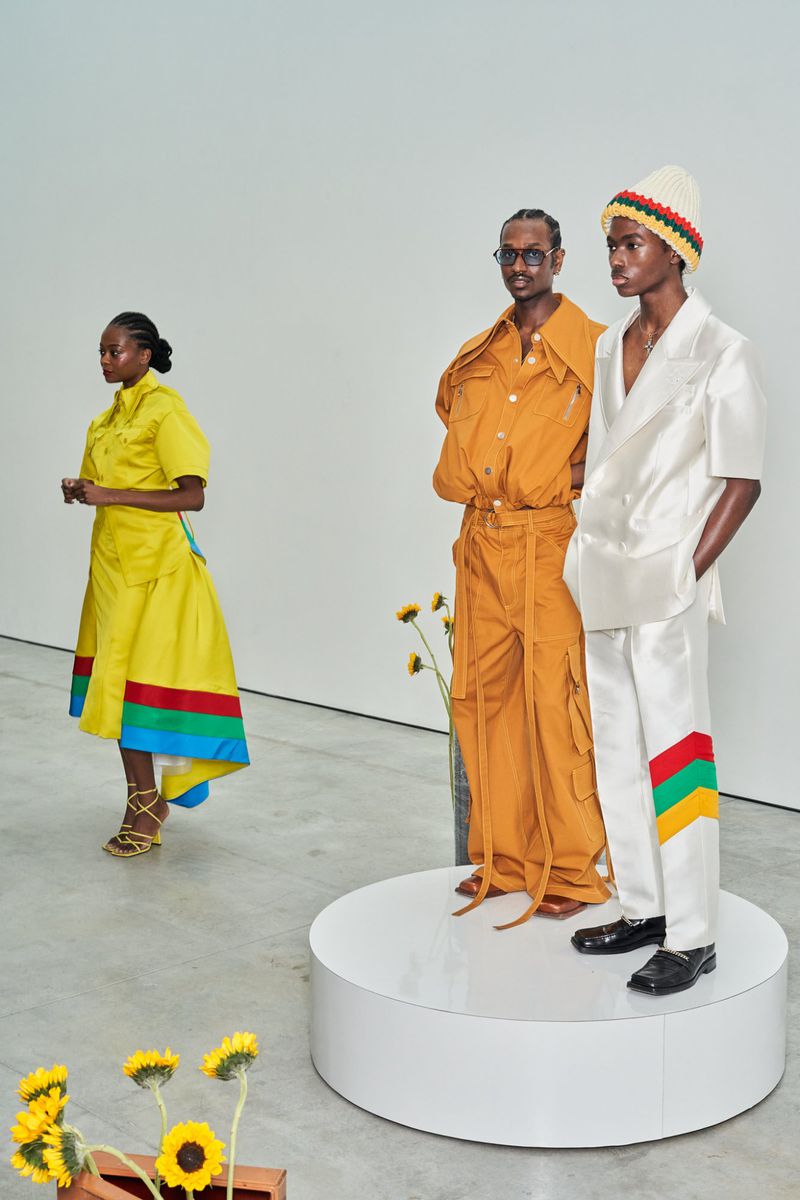 Three models showcase fashion designs on a white platform and in the background, featuring yellow, orange and white outfits with embellishments.