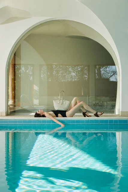 Woman in black swimsuit relaxing by a turquoise pool next to a white arched poolside building with a woven bag on the edge.