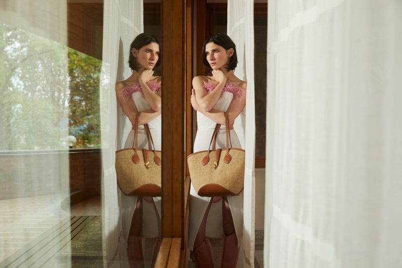 Woman in white dress holds straw tote bag, posing by window with curtains. Natural light, soft colors, fashion shot.