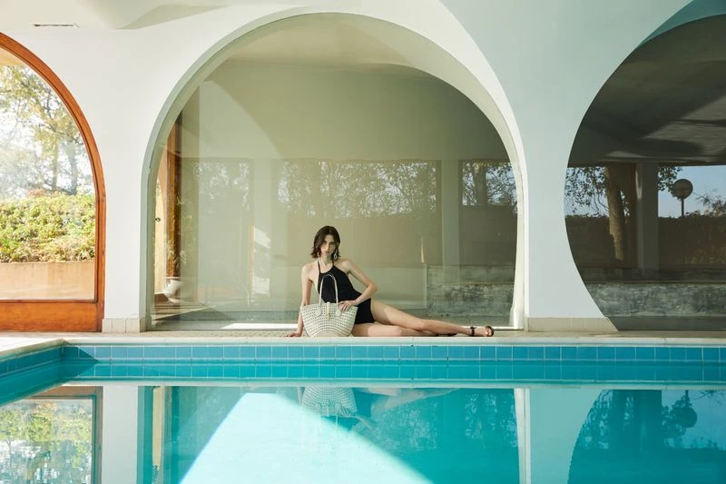 Brunette model in black swimsuit poses near pool with woven bag, reflecting serene indoor leisure and fashion.