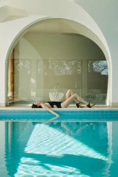 Woman lying beside a swimming pool with a white woven handbag resting on her torso against an arched window.