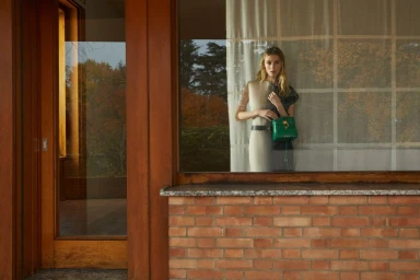Woman holding a green handbag standing behind a window in a brick and wood architecture setting