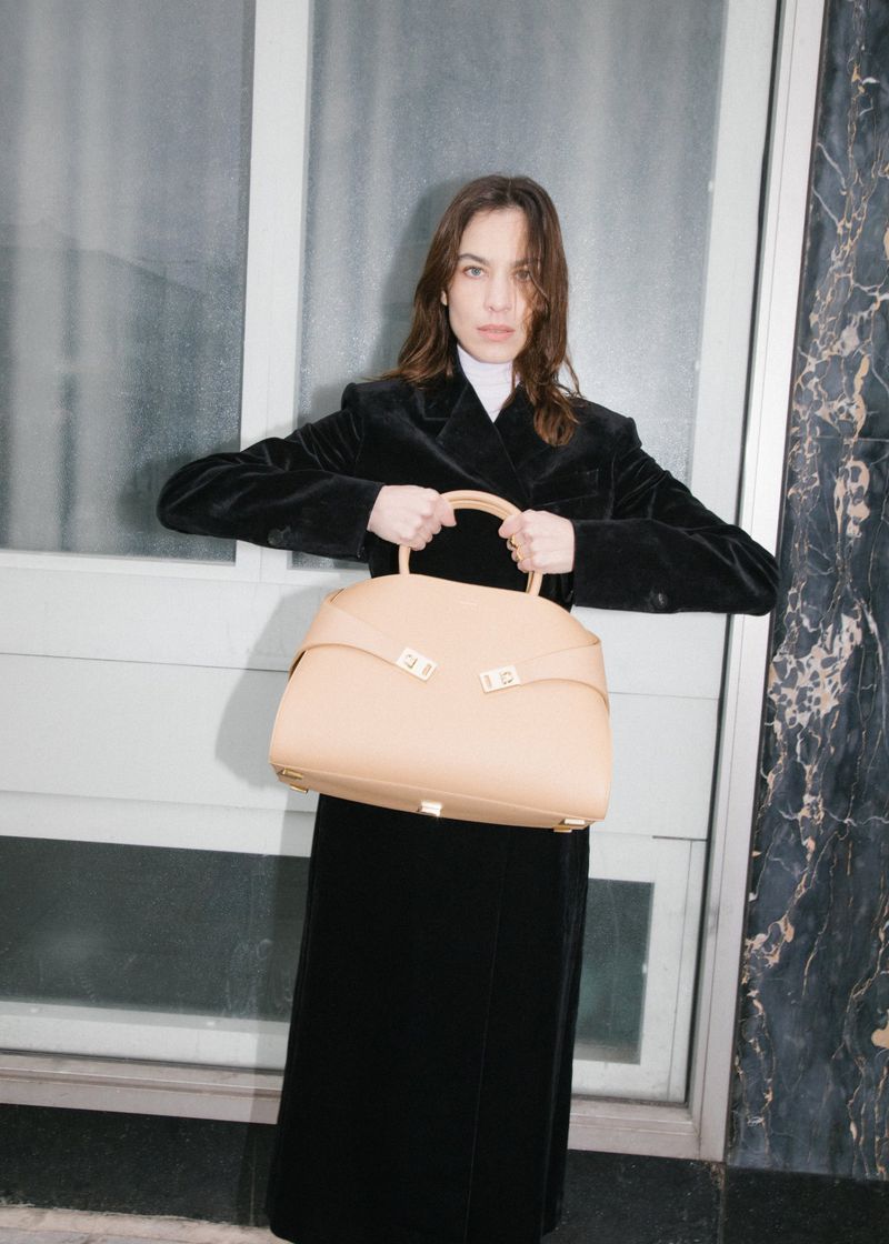 Elegant woman in a black velvet coat holds a large tan leather handbag against a neutral background in a fashion forward pose.