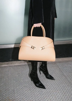 Person holding a structured beige leather handbag, wearing a black coat and glossy black heeled boots on textured metal flooring