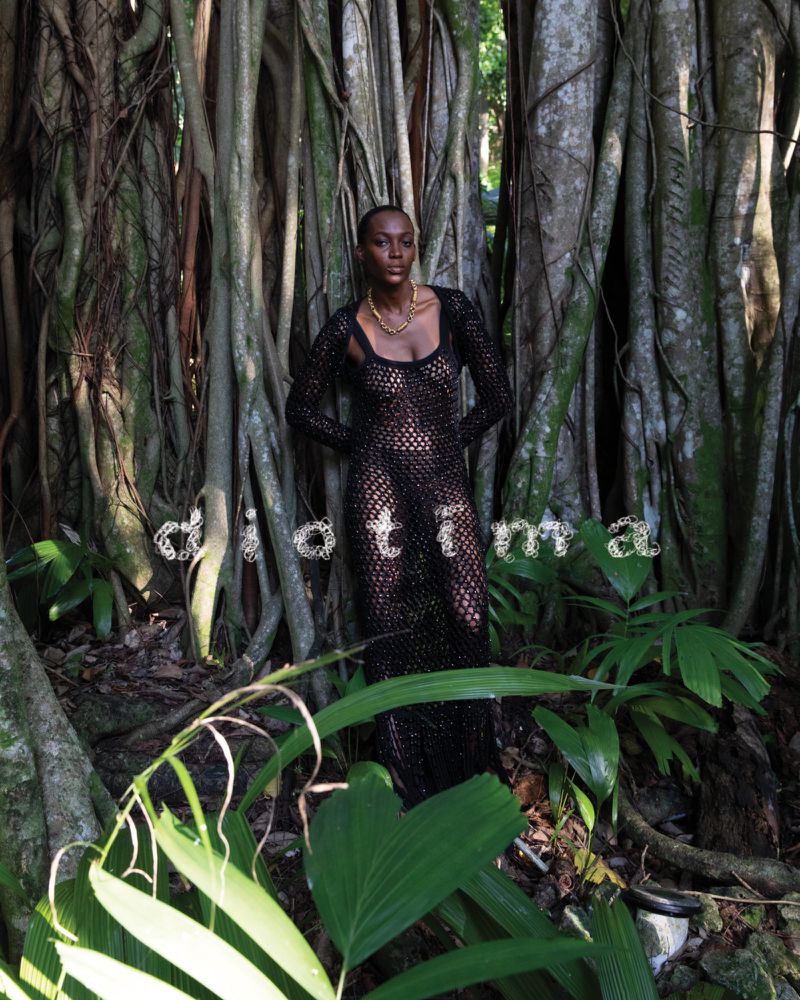 An adult woman poses in a sheer, black dress amongst lush jungle greenery with large trees and bright, green leaves in an outdoor setting.
