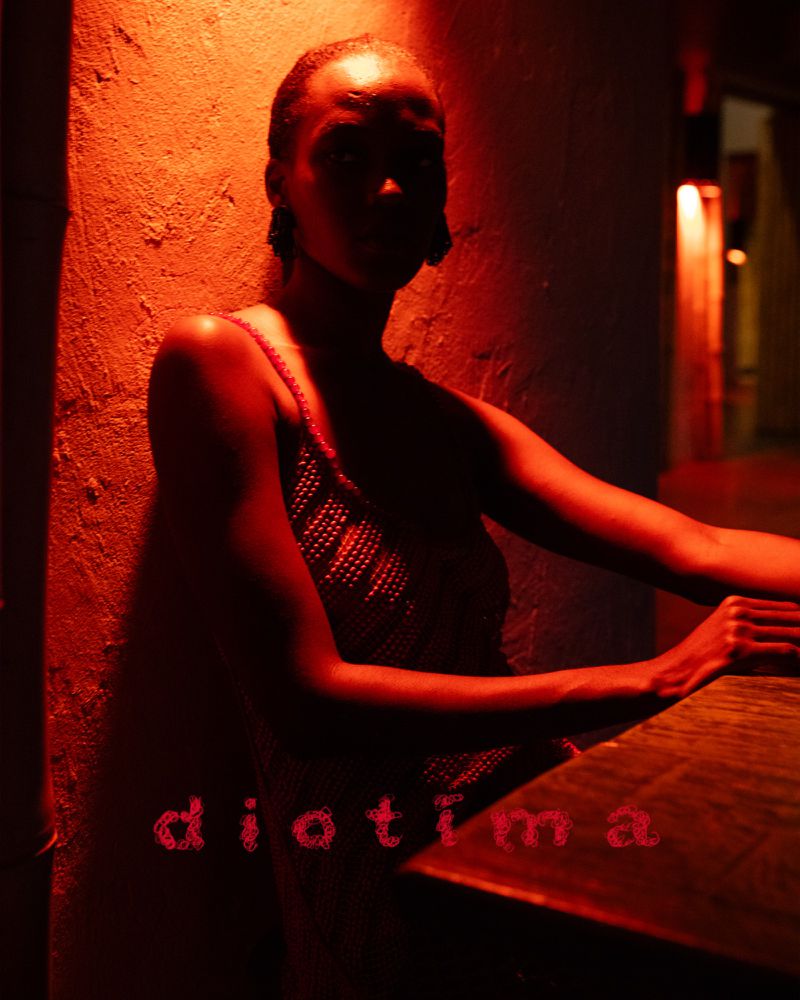 Portrait of adult female with dark hair wearing a mesh tank top leaning against a wall in a dimly lit indoor setting with red lighting.