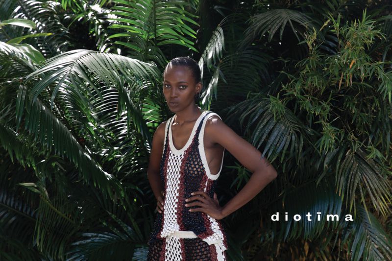 A woman with dark skin poses in a crocheted dress against a backdrop of lush green palm trees, creating a natural and fashionable scene.