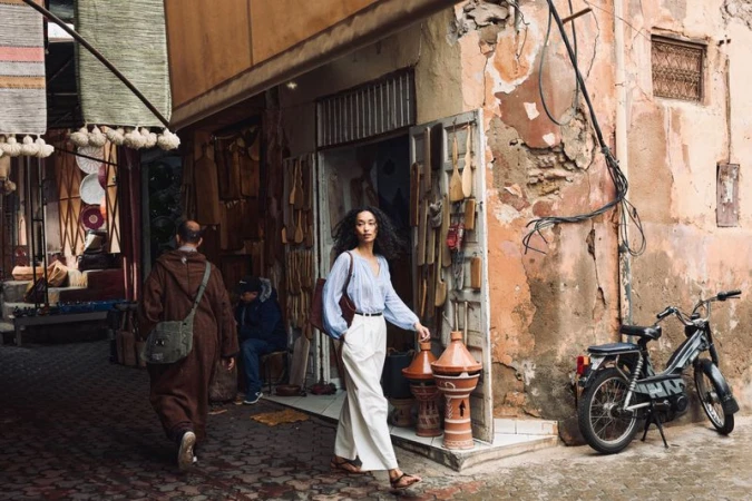 Fashionable woman in blue blouse and white pants strolls through a vibrant Moroccan street market with detailed architecture and nearby scooter.