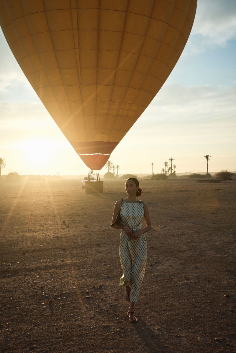 Woman in patterned jumpsuit walks toward the camera on a desert landscape with a hot air balloon in the background at sunrise.