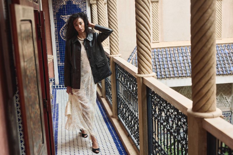 A woman in a dress and jacket poses on a tiled balcony with carved columns. She holds a small bag and wears heels in a fashion editorial.