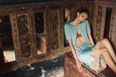 Anais Mali wearing a light blue knit cardigan and lace-trimmed satin skirt, reclining against an ornate vintage wooden mirror panel.