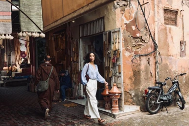 Model walking along a street market wearing a light blue blouse and high-waisted white wide-leg trousers past an artisan shop.