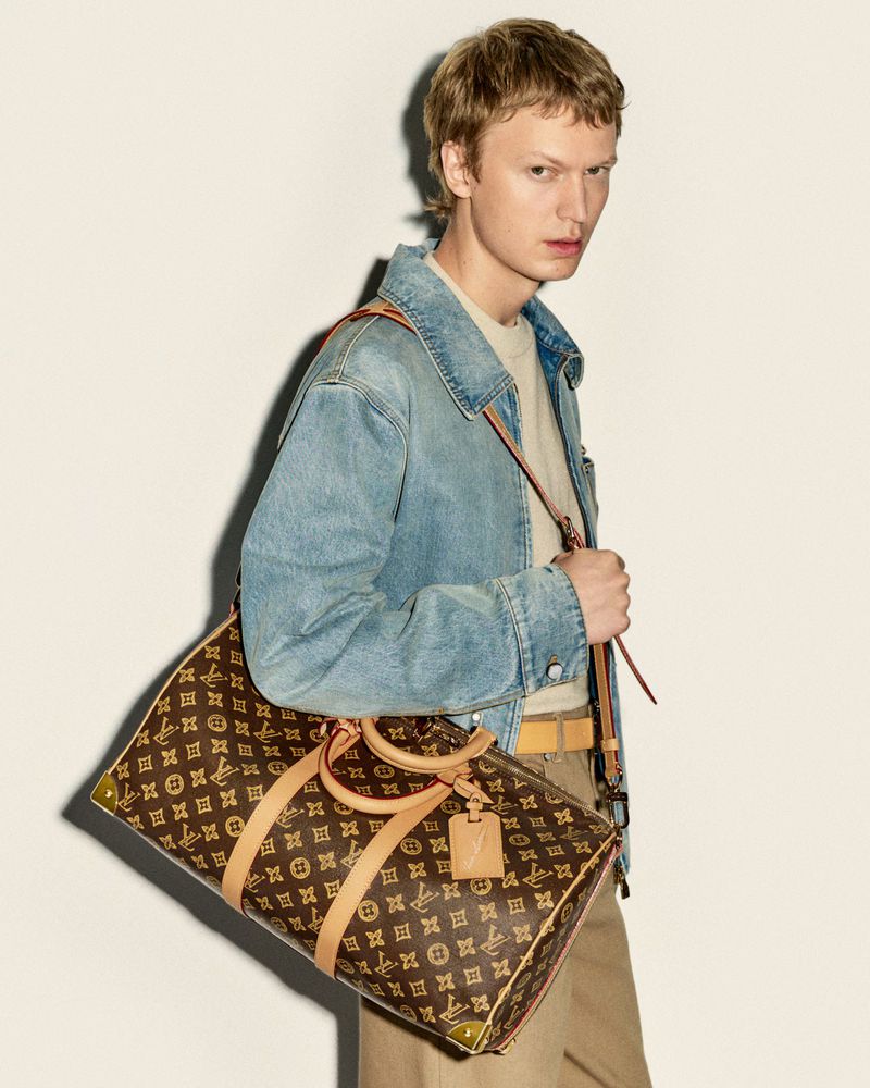 Man wearing denim jacket and khakis with Louis Vuitton duffel bag in a studio setting against a neutral wall.