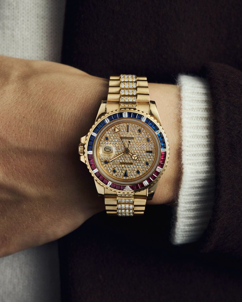 Close-up of a person's wrist wearing an ornate gold wristwatch with ruby and sapphire accents. Gold bracelet and clothing visible.