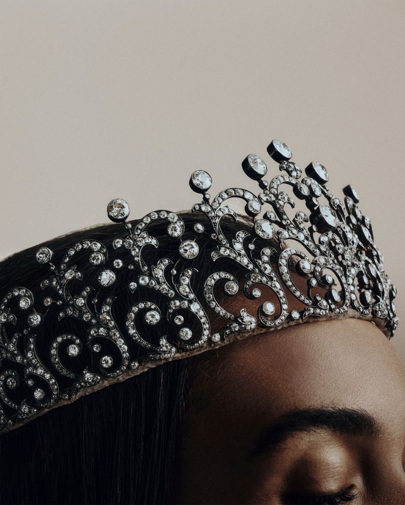 Close-up of a dark-haired adult female wearing an ornate silver-and-diamond crown against a pale background, creating a regal and elegant image.