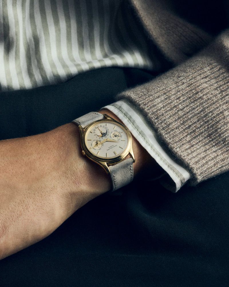 Close-up of a fair-skinned arm wearing a gold watch with a light gray leather band, a beige sweater, and a white striped shirt.