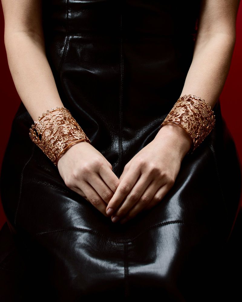 Close-up studio shot of a person wearing large copper bracelets, seated with hands clasped over shiny black leather attire against a red background.