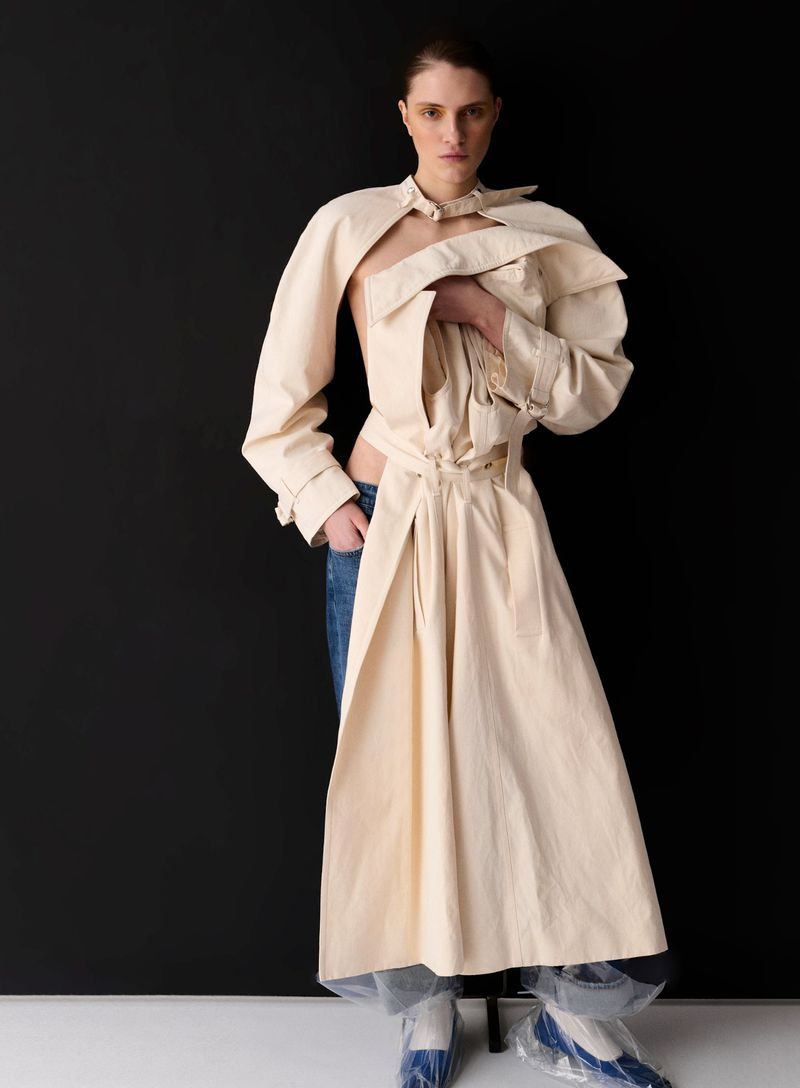Fashion shot of a woman in a beige trench coat and blue jeans against a black background in a photo studio setting. Medium shot.