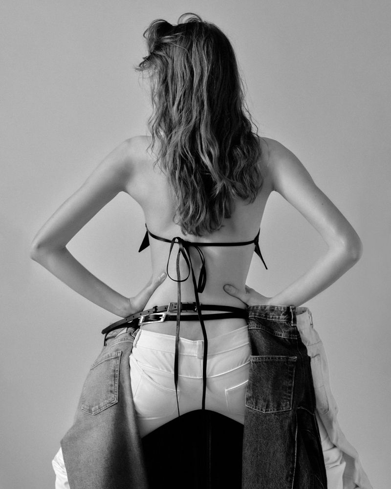 Back view of a slender woman with long wavy hair wearing a bra top and white pants. Jeans hang at her waist. Black and white studio shot.