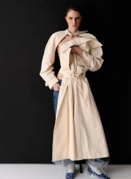 Model Alice Cooper wearing a deconstructed cream trench coat and blue jeans against a dark studio background.