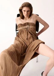 Model Alice Cooper wearing a pleated strapless brown dress with a wide belt across the bodice, seated against a white background.