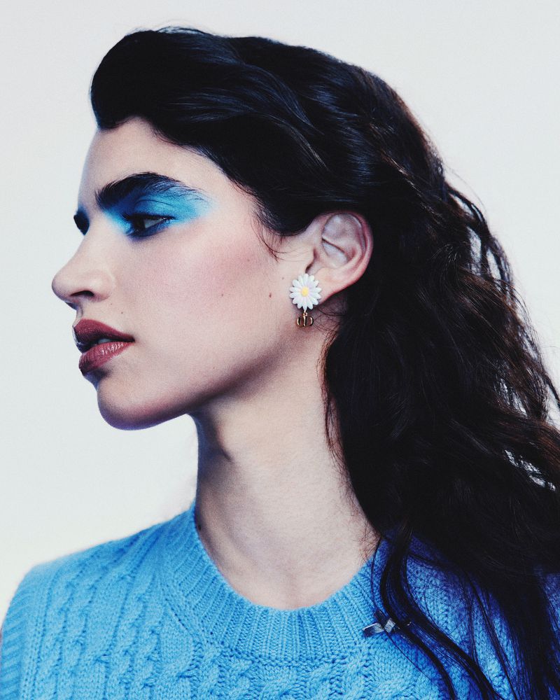 Profile of an adult woman with dark hair wearing a cable-knit sweater, bright blue eyeshadow, and daisy earrings against a white backdrop.