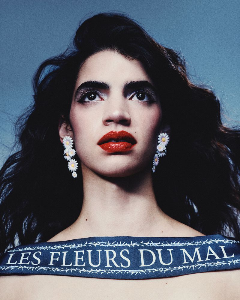 A young adult woman with dark hair and red lipstick gazes upward, wearing daisy earrings and a blue ribbon necklace with white text.