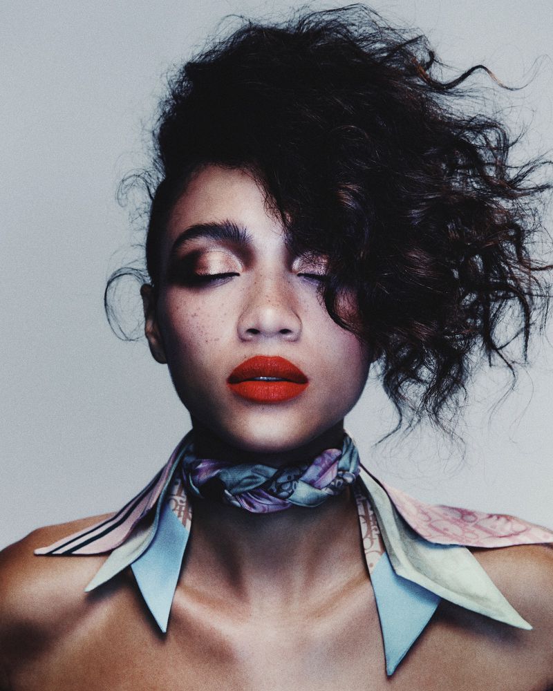 Portrait of a young woman with striking red lipstick, eyes closed, wearing a patterned scarf around her neck against a neutral background.