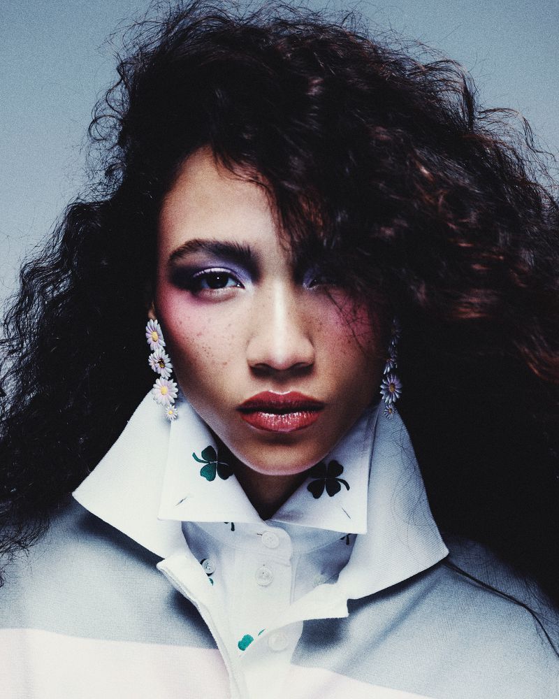 Close-up shot of a woman with curly hair and bold makeup wearing a white rugby shirt with clover motifs and daisy earrings.