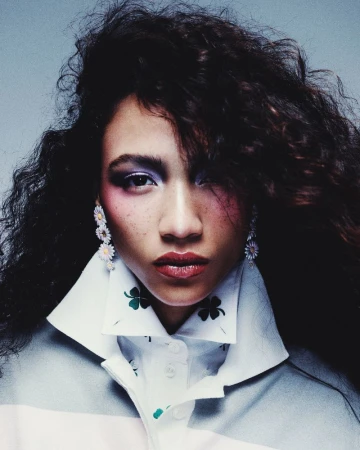 Close-up shot of a woman with curly hair and bold makeup wearing a white rugby shirt with clover motifs and daisy earrings.