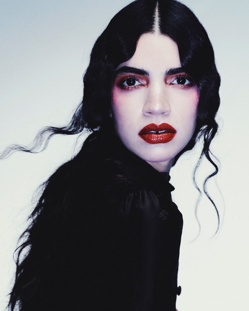 Portrait of a woman with dark, wavy hair and red lipstick, wearing a high-necked dark top, against a light grey studio background.