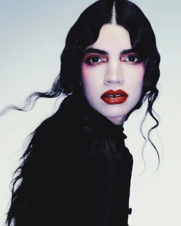 Portrait of a woman with dark, wavy hair and red lipstick, wearing a high-necked dark top, against a light grey studio background.