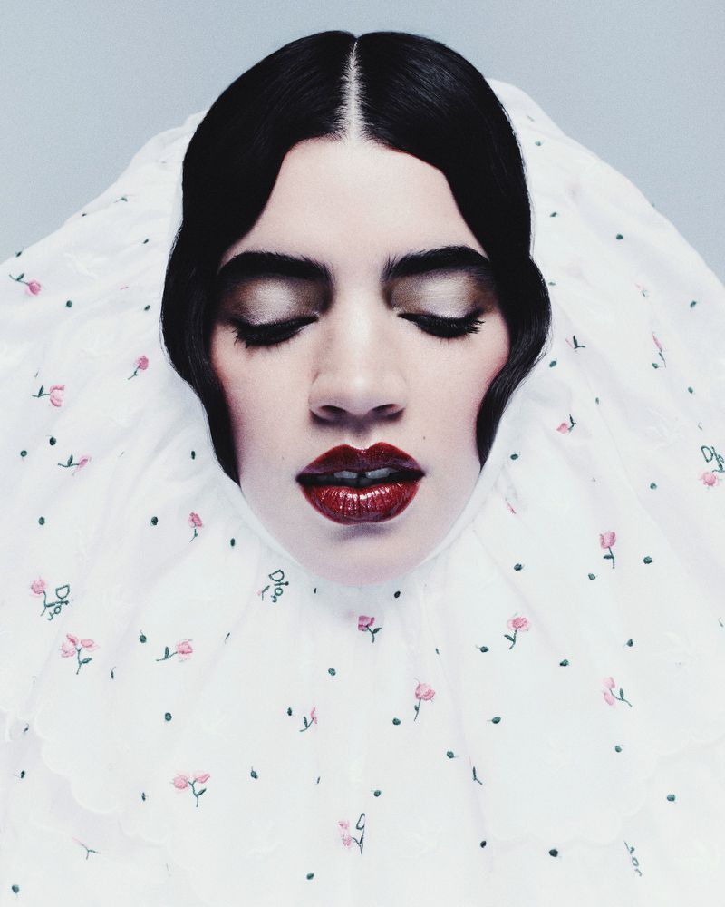 Headshot portrait of a serene woman with closed eyes, dark hair, and red lipstick. She is framed by a white floral-patterned garment.